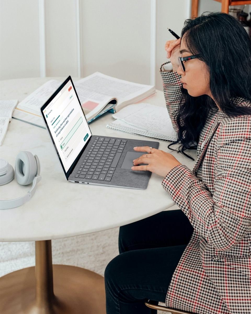 A student in glasses taking an RBT practice quiz on her laptop, with an open textbook and handwritten notes on the table